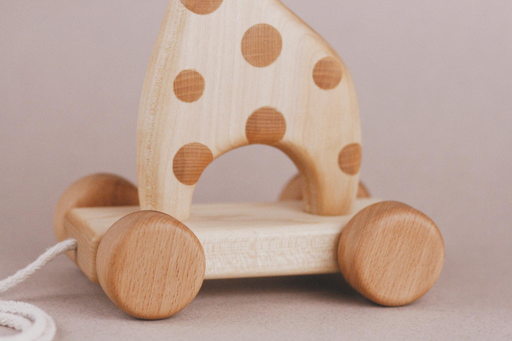 The Handmade Wooden Giraffe Pull Toy features rounded wheels and a playful pull string. Its body is adorned with circular brown spots and sits charmingly on a light beige surface, making it an enchanting toy for children.