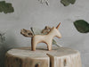 A small, Handmade Wooden Unicorn carved from maple wood with a yarn mane and tail stands on a tree stump. The background is a gray wall adorned with green leaf decorations and a paper snowflake. Ideal for children 3 and up, the scene has a rustic and whimsical feel.