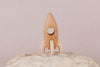 A Handmade Wooden Rocket crafted from beech wood, complete with a circular window, stands gracefully on a textured rock surface against a plain backdrop. It boasts smooth, natural wood tones accentuated by linseed oil and features simple detailing.