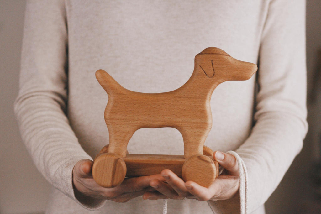 A person in a beige sweater holds the Handmade Wooden Dog Pull Toy, crafted from smooth maple and beech wood and featuring simple carved details along with four wheels.