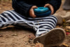 A toddler, wearing striped pants and sneakers, sits on the ground holding a Rainbow Wooden Stacker with a beeswax finish, with autumn leaves scattered around. Only the child's lower body and hands are visible.