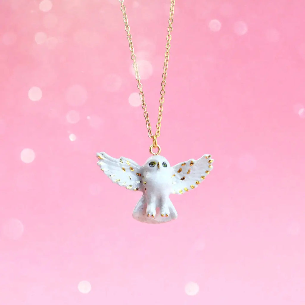 A Gold and White Snowy Owl Necklace with a pendant shaped like an owl with wings spread wide, adorned with 24k gold-plated accents. The background is a soft, bokeh pink gradient, creating a dreamy and whimsical atmosphere.