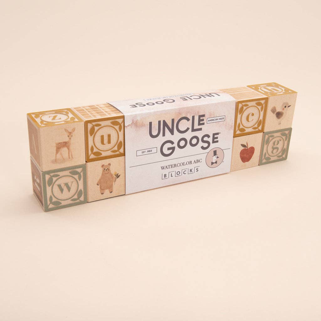 A rectangular box of Uncle Goose Watercolor ABC Blocks is displayed, made from sustainable basswood. The packaging showcases a paintbox theme adorned with images of animals, an apple, and letters in soft pastel hues.