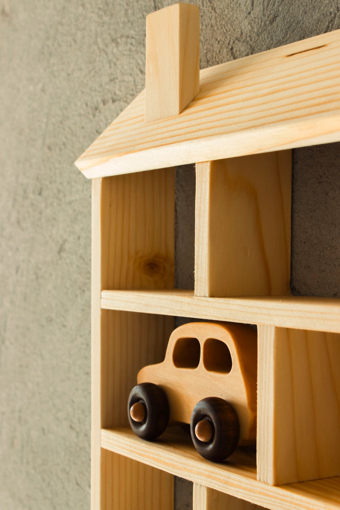 A Wooden Shelf No. 22 designed to look like a house with a roof and chimney is mounted on a textured wall. Ideal for kids' storage shelves, it features a small, simple wooden toy car on the middle shelf, creating a cozy and nostalgic aesthetic perfect for any children's room.