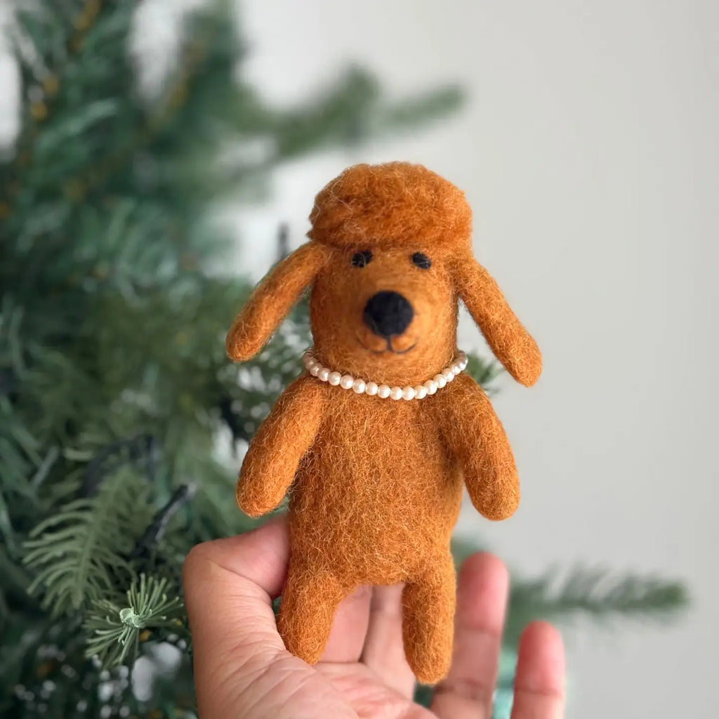 A person holds up a small, hand-stitched Poodle Dog Finger Puppet against a blurred background of a pine tree. The poodle is brown with floppy ears and a round black nose, and it is wearing a tiny pearl necklace around its neck.