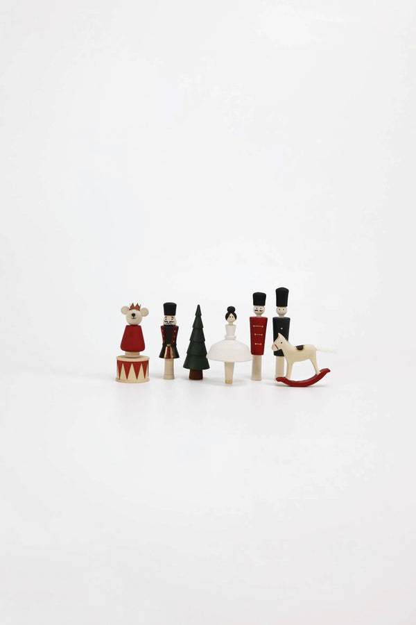 Against a plain white background stands a delightful set of Wooden Nutcracker Figurines handcrafted from linden wood. The collection includes a bear, soldiers harking back to classic Nutcrackers, a tree, and a sled. These whimsical wooden toys are dressed in striking colors of red, white, green, and black that beautifully capture their artistic charm.