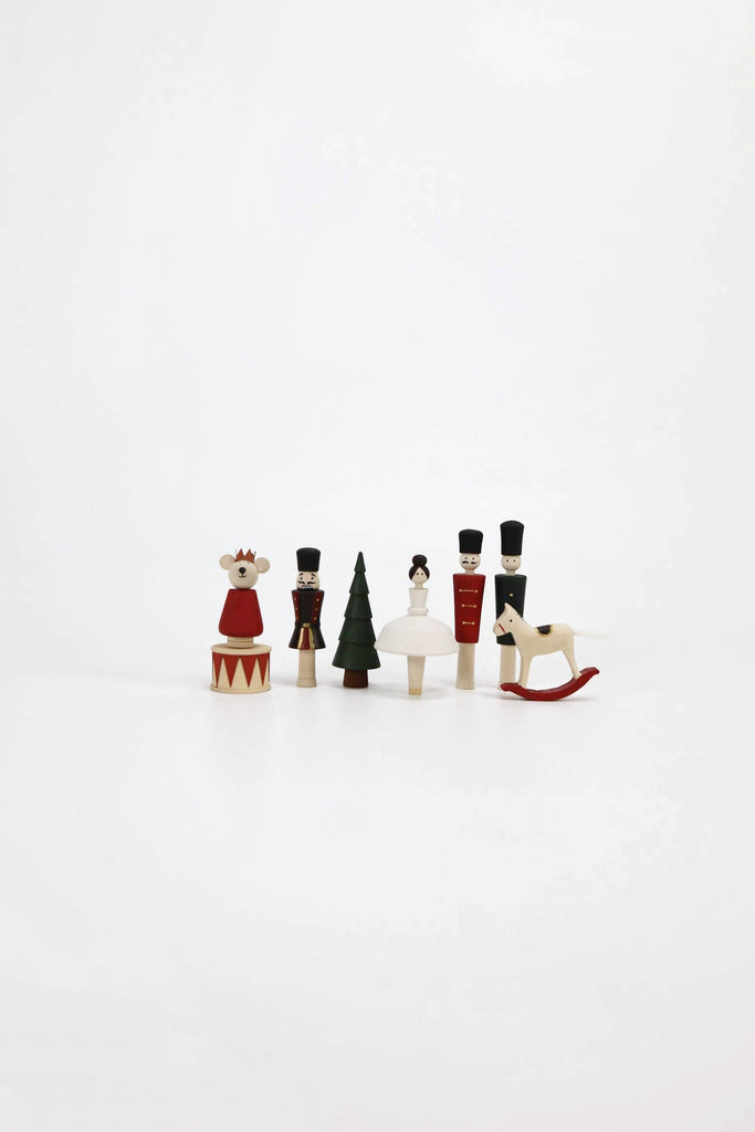 Against a plain white background stands a delightful set of Wooden Nutcracker Figurines handcrafted from linden wood. The collection includes a bear, soldiers harking back to classic Nutcrackers, a tree, and a sled. These whimsical wooden toys are dressed in striking colors of red, white, green, and black that beautifully capture their artistic charm.