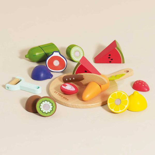 A vibrant collection of toy fruits and vegetables, featuring watermelon, kiwi, and lime, alongside a chopping board set and toy peeler, is displayed on a beige surface. The Wooden Chopping Board & Sliceable Play Food is perfect for role play fun while enhancing fine motor skills.