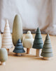 The Wooden Forest Set from SABO concept toys features a charming arrangement of small, stylized wooden trees in diverse shapes and shades of green and cream, displayed on a light brown surface. The softly blurred background creates a cozy and artistic atmosphere, ideal for imaginative play.