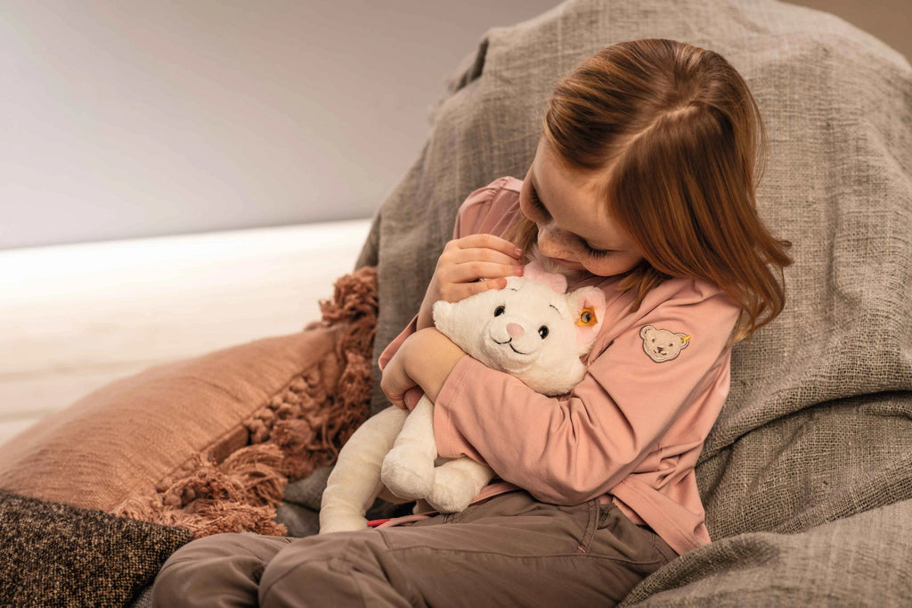 A young girl with light brown hair, wearing a long-sleeved pink shirt and beige pants, is sitting and hugging a Steiff, Disney's "Aristocats" Marie Cat Plush Stuffed Toy, 9 inches. She is looking down at the toy with a gentle and affectionate expression. The background is neutral and softly lit.