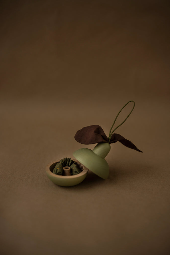 The "Wooden Pear & Little Friend" rests on a brown surface, characterized by its small, green, bell-shaped container adorned with a fabric leaf and stem. The container's top opens with a magnetic closure to reveal intricate green contents wrapped in fabric inside. A simple matching brown background enhances the arrangement with an earthy tone.