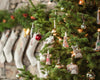 A decorated Christmas tree, lovingly adorned with Fox Tree Trimmer Ornaments and shiny baubles, stands in a cozy room, sparking Christmas excitement. In the background, white stockings hang on a stone wall, evoking cherished holiday memories.