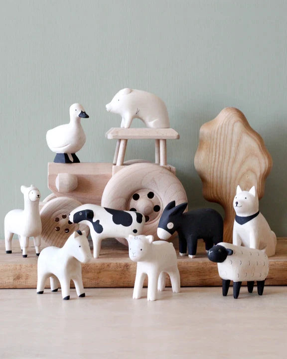 A collection of handcrafted wooden farm animal toys, meticulously crafted by Bali artisans using Albizia wood, includes a cow, donkey, horse, sheep, dog, pig, goose, and llama. Each toy is painted in minimalistic black and white and arranged elegantly in front of a wooden tree and tractor. The collection is called Handmade Tiny Wooden Farm Animals - Akita Dog.