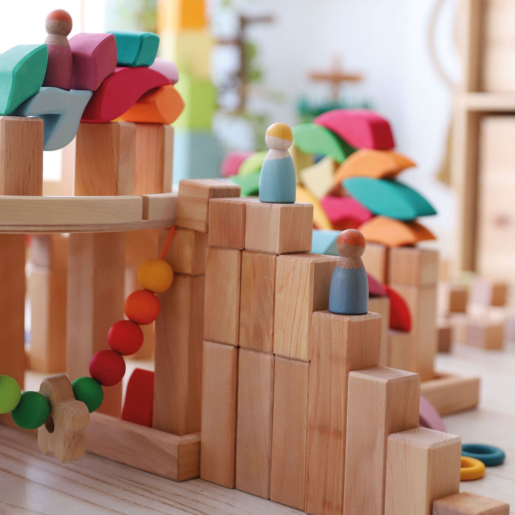 A colorful wooden toy set featuring block structures, bridges, and peg dolls arranged in a whimsical manner. This setup includes 100 building blocks and resembles a playful landscape on a wooden surface. The natural wood blocks and brightly painted elements create an enchanting scene with Grimm's Large Natural Stepped Pyramid.