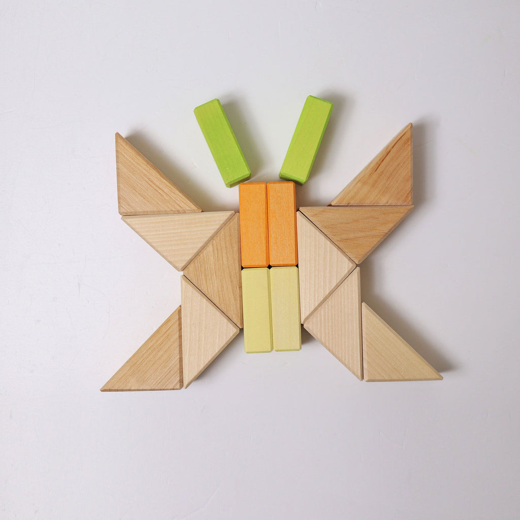 A butterfly shape composed of geometric shapes arranged on a white surface using the Pastel Duo Block Set. The wings are crafted from triangular and rectangular building blocks in natural wood tones, while the body features yellow and orange blocks, and the antennae are made from green blocks.