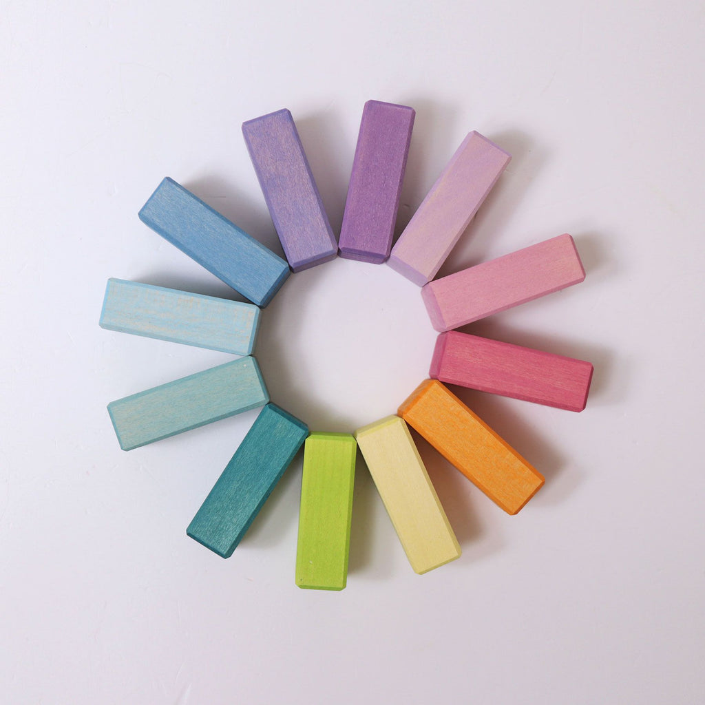 A circular arrangement of twelve rectangular wooden blocks from the Pastel Duo Block Set features an array of vibrant shades, including purple, pink, red, orange, yellow, green, and blue. These geometric shapes are evenly spaced on a white background, forming a visually pleasing rainbow gradient.