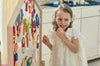 A young girl wearing a white dress decorated with daisy patterns stands next to a NORITER Signature Double-Sided Board Bundle, which is covered in vibrant shapes and drawings. She smiles brightly and gazes to the side, appearing engaged and cheerful in the well-lit room.