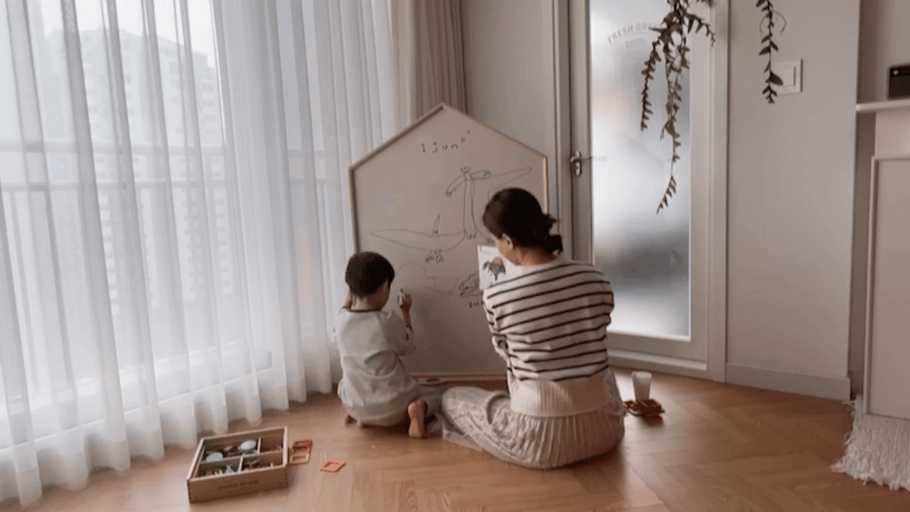 A child and an adult sit on the floor, drawing on a NORITER Signature Double-Sided Board Bundle in a room with soft natural light. Toys are scattered nearby, and plants hang in the background, creating a cozy atmosphere as colorful Fruit Felt pieces add charm to their creative space.