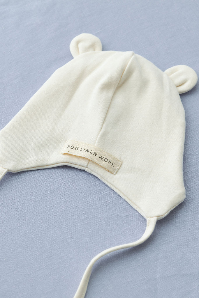 A Baby Pilot Hat with bear ears, designed for 0-3 months, lying flat on a light gray surface. Made from organic cotton, the hat has a tag that reads "Fog Linen Work" and features two strings for tying under the chin.