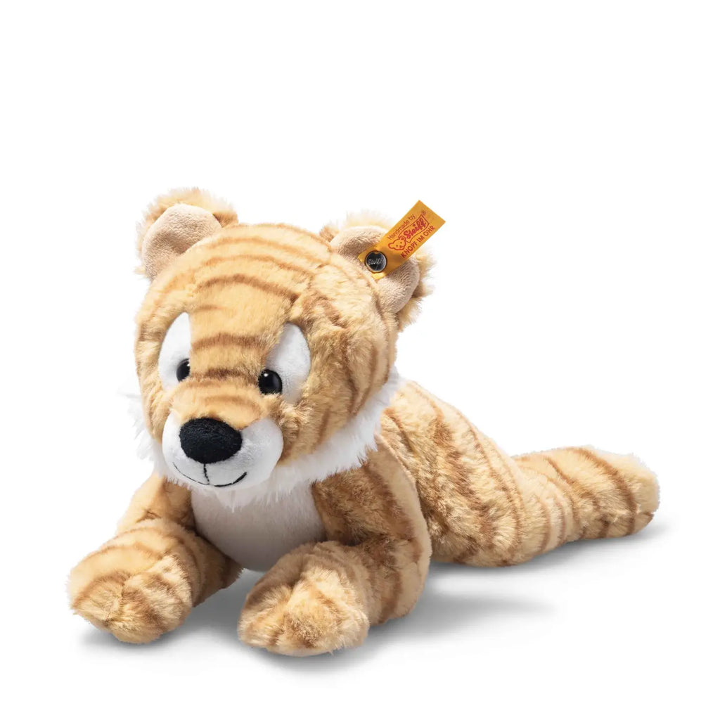 A Steiff, Toni Tiger Stuffed Animal Plush, 12 Inches with a light brown and beige striped coat, large round eyes, and white facial features is lying down on its stomach with front paws extended forward. A small yellow tag is attached to its left ear.