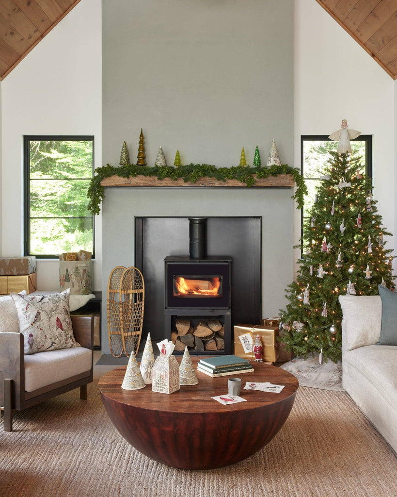 The living room exudes coziness with a wood-burning stove nestled in the fireplace, beautifully decorated with small Christmas trees and garlands featuring metallic threads. A large tree stands festively adorned, charmingly accented by an Angel Dog Ornament. Rustic wooden coffee tables and comfy sofas contribute to the warm ambiance, while expansive windows offer a view of the lush green landscape outside.