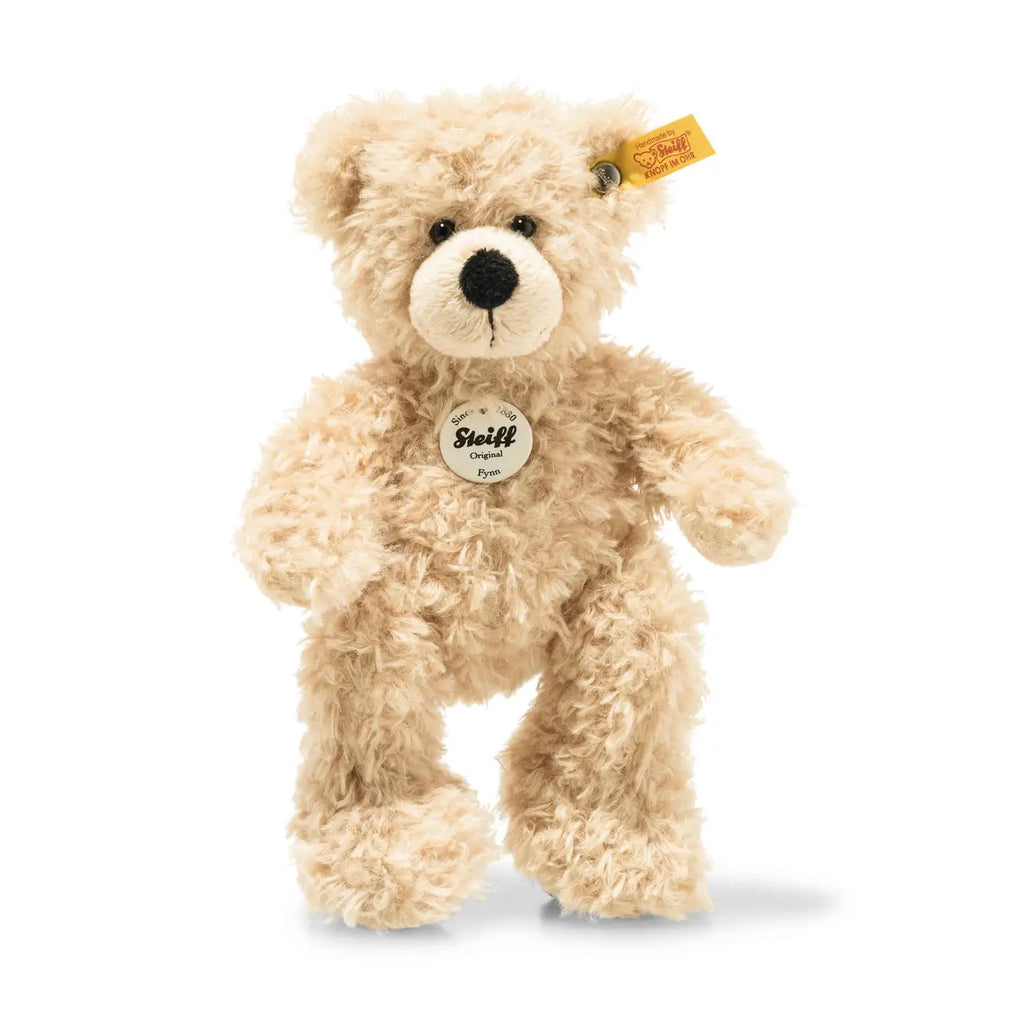 A Steiff, Fynn Teddy Bear Plush Toy, 7 Inches with a Steiff tag on its ear and an emblem on its chest. The bear, known for its "Button in Ear" trademark, has a black nose and eyes, featuring a plush, shaggy texture and standing upright on its hind legs. This exquisite handmade plush is a timeless treasure.