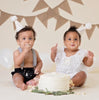 Two babies, each sporting a White Party Hat adorned with a "1," are seated on the floor delighting in a white frosted cake. Their faces and outfits are covered in cake, beautifully encapsulating the joy of birthday celebrations. The setting features white balloons and a beige bunting banner in the background.