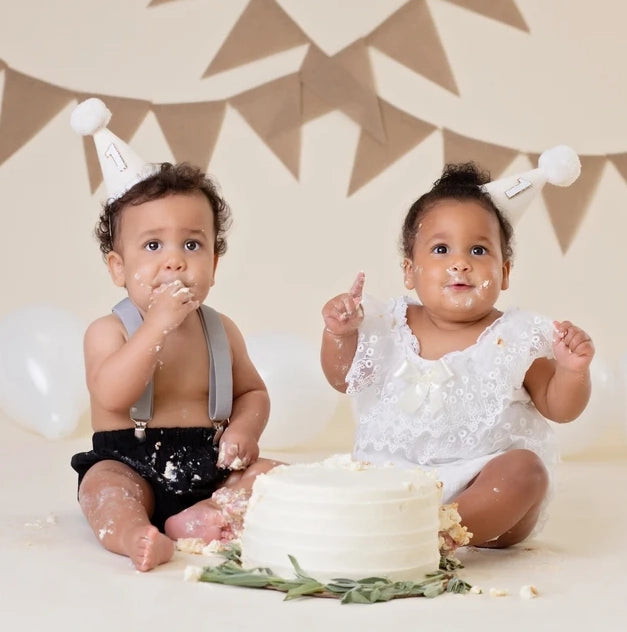 Two babies, each sporting a White Party Hat adorned with a "1," are seated on the floor delighting in a white frosted cake. Their faces and outfits are covered in cake, beautifully encapsulating the joy of birthday celebrations. The setting features white balloons and a beige bunting banner in the background.