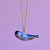 A delicate gold chain necklace featuring a small, hand-painted blue whale pendant against a soft purple background. The Blue Whale Necklace, adorned in aquamarine tones with a glossy finish, captures the whimsical nature of the sea creature.