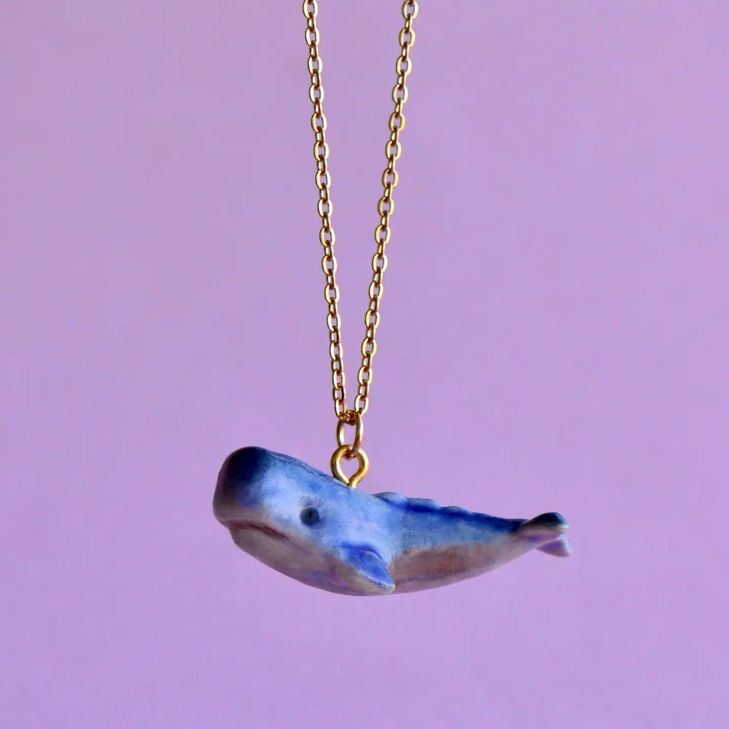 A delicate gold chain necklace featuring a small, hand-painted blue whale pendant against a soft purple background. The Blue Whale Necklace, adorned in aquamarine tones with a glossy finish, captures the whimsical nature of the sea creature.