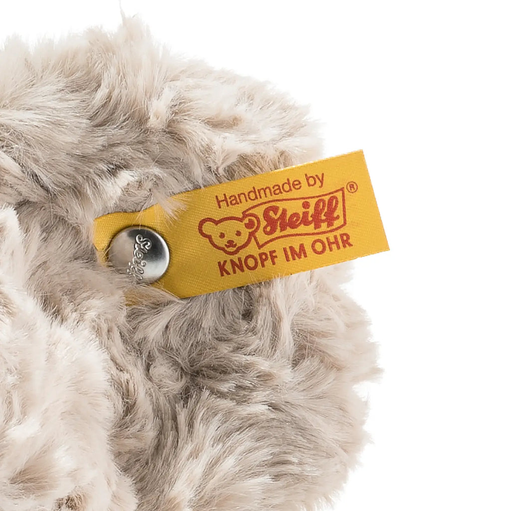 Detailed shot of the Steiff, Honey Teddy Bear's fur showcasing a yellow fabric tag. The tag highlights a logo featuring the iconic face of the Honey Teddy Bear and includes German text: "Handmade by Steiff KNOF IM OHR." A metal button, elegantly embossed with script, is securely attached to the tag. This beloved 15-inch plush toy can also be conveniently machine washed.