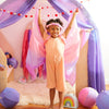 A young child stands joyfully with arms raised under a play canopy draped with pink and purple fabric, decorated with heart garlands. They are engaged in dress-up play, wearing an orange outfit, a Sarah's Silk Blossom Garland Headband, and surrounded by colorful toys and cushions.