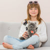 A young girl with blonde hair sits cross-legged on a light gray sofa. She is wearing a white sweater with cat designs, light blue jeans, and pink sunglasses on her head. The child-friendly girl is holding a Teddy Hermann Gray Sitting Cat Stuffed Animal with a tag on its neck, and she smiles at the camera.