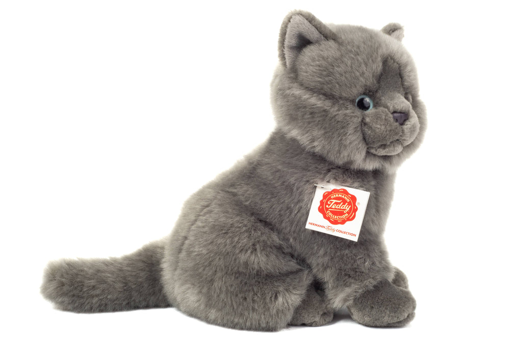 The Teddy Hermann Gray Sitting Cat Stuffed Animal features a soft, fluffy texture and blue eyes. This child-friendly, high-quality plush toy sits upright and has a tag on its neck with an orange label that reads "Teddy" along with some additional text. Designed to look realistic, the cat also has a bushy tail.