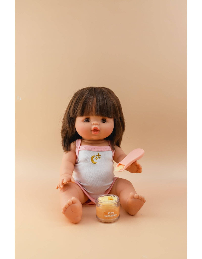 A doll with brown hair sits against a beige background, dressed in a light-colored sleeveless outfit featuring a crescent moon and star. It holds a pink spatula and is accompanied by a small jar labeled "Tiny Tummies - Peach jelly food," ideal for conjuring magical spoonfuls of doll food delights.