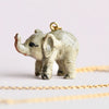 A small, intricately detailed Baby Elephant Necklace hand-painted and crafted from a beige material with black accents, featuring a raised trunk and 24k gold plated chain. The background is a soft, light purple, highlighting the pendant’s majestic beauty and delicate craftsmanship.