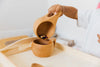 A child pours beans from a Wooden Pouring Cup into a bowl on a tray, focusing on Montessori-style practical life activity using early learning tools for sensory and motor skills development.