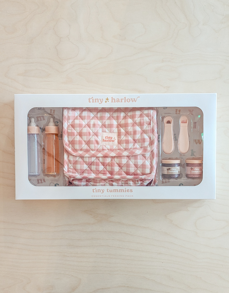 A delightful "Tiny Tummies Pink Gift Box - The Essentials" by Tiny Harlow sits on a wooden surface, showcasing a checkered pouch along with doll essentials such as two small bottles, utensils, and tiny jars—all components of the tiny tummies essential feeding pack—perfect for nurturing little imaginations.