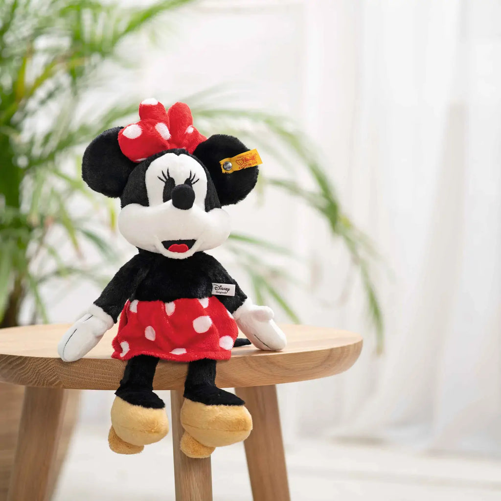 The Steiff, Disney's Minnie Mouse Stuffed Plush Toy, 12 Inches, from the Soft Cuddly Friends Collection, is pictured sitting on a wooden stool indoors. The toy is dressed in a red polka dot dress with a matching bow and yellow shoes. The background features a blurred green plant and white curtains.