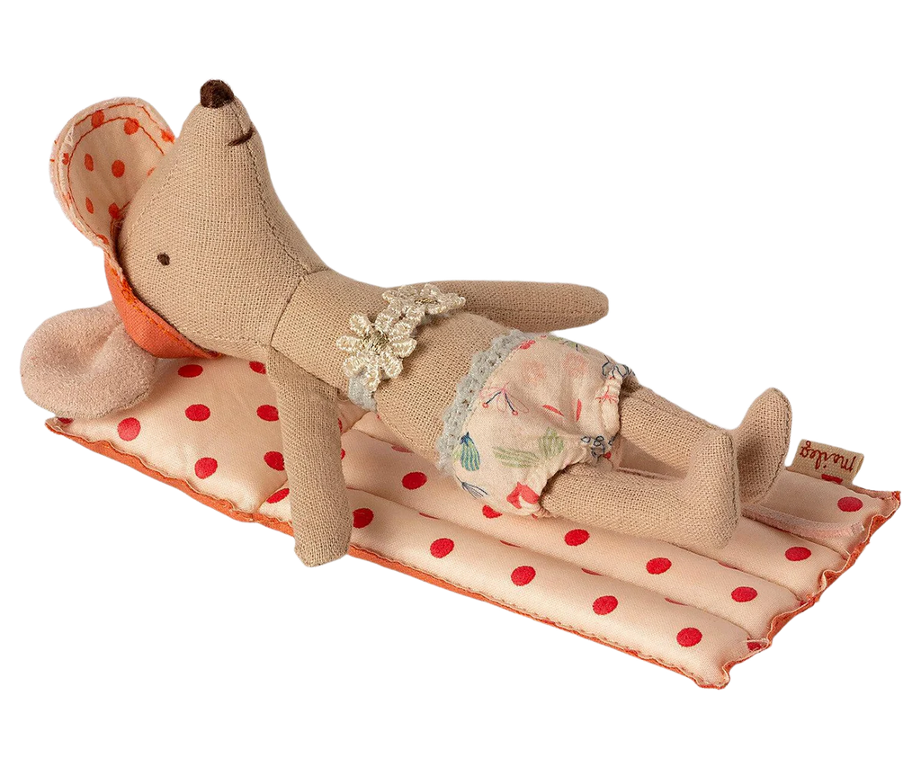 A small fabric mouse doll from Maileg Beach Float, Mouse - Red dot, lies on a polka-dotted beach mat. It is wearing a big sun hat, floral shorts, and has a lace accessory around its neck. The doll's beige-coated fabric body is stuffed and appears relaxed as if sunbathing.