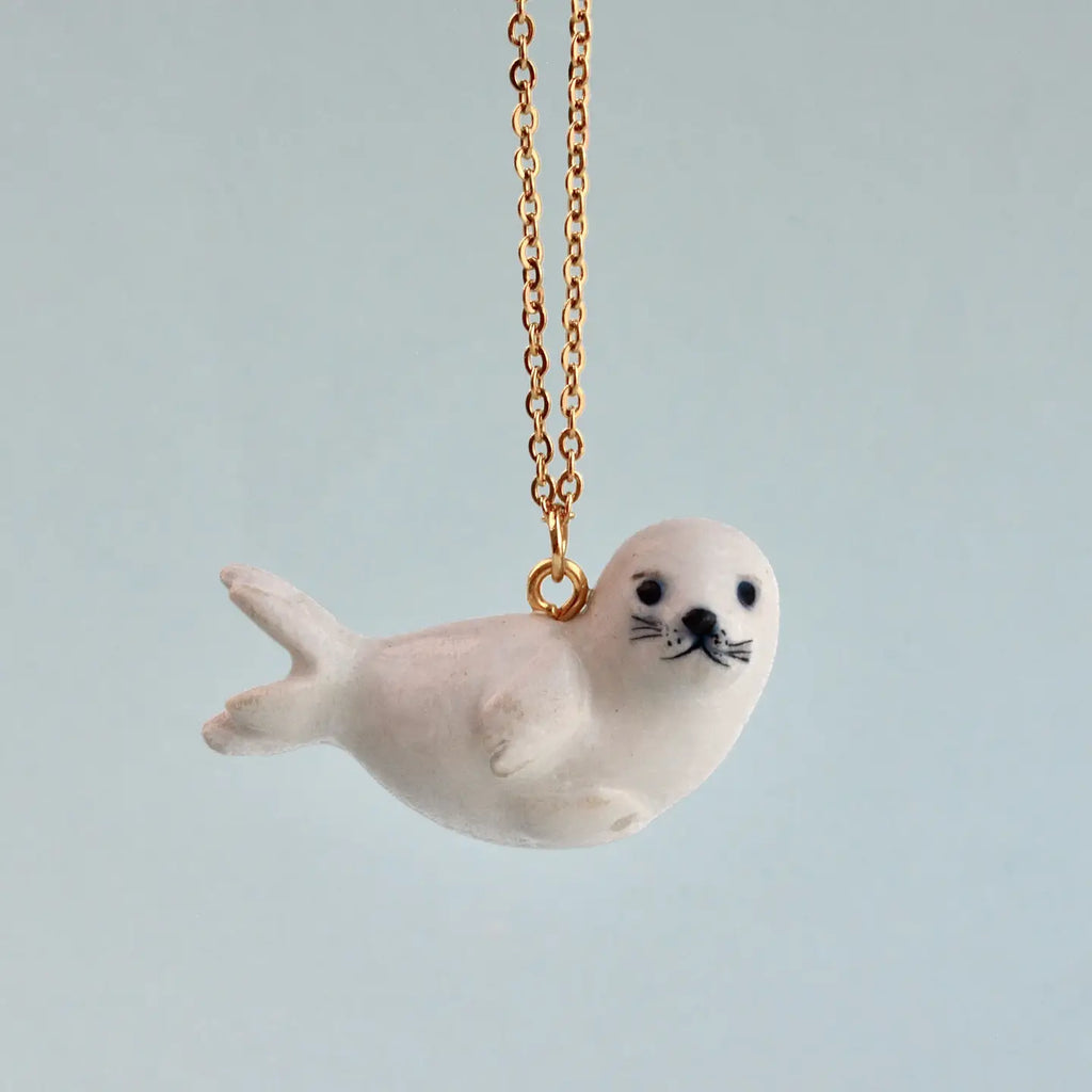 A small, white Seal Necklace hangs from a delicate 24k gold plated chain against a light blue background. The hand painted jewelry piece has black eyes and whiskers, giving it a cute and whimsical appearance.