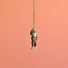 A Sasquatch Necklace hangs from a gold chain necklace against a peach-colored background. The handcrafted jewelry piece features the monkey holding onto the chain with one hand, appearing playful and animated. This limited-edition fashion accessory adds a touch of whimsy to any outfit.