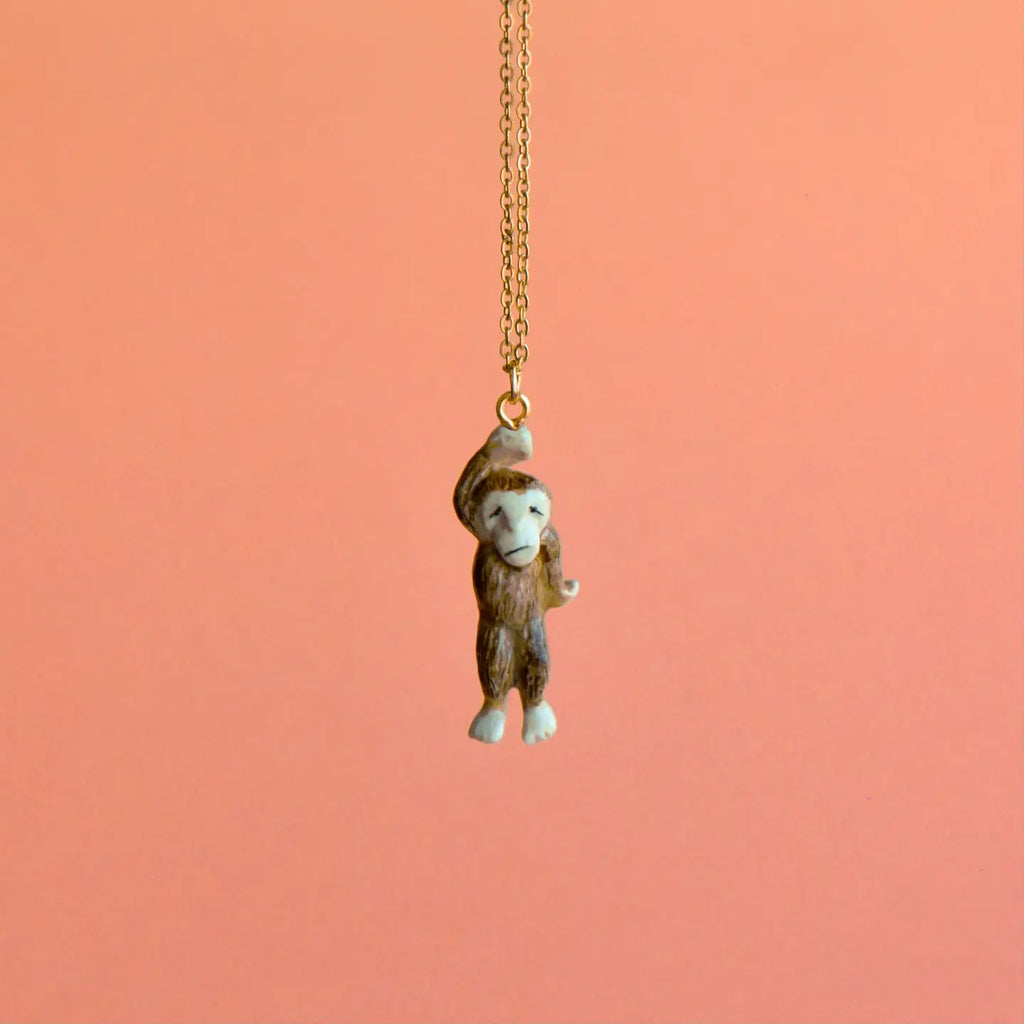 A Sasquatch Necklace hangs from a gold chain necklace against a peach-colored background. The handcrafted jewelry piece features the monkey holding onto the chain with one hand, appearing playful and animated. This limited-edition fashion accessory adds a touch of whimsy to any outfit.