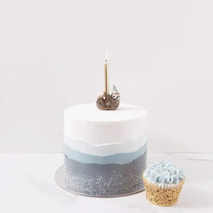 A two-tiered cake with blue and white ombre layers, decorated with a gold candle and a small, hand-painted Sloth Cake Topper, next to a cupcake with blue frosting on a