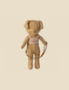 A well-worn vintage teddy bear stands upright, wearing a velvety harness that spells out "Maileg Puppy Supply, Harness" in cursive. The bear is fuzzy, with a light brown color and floppy ears. The background is light beige and empty.