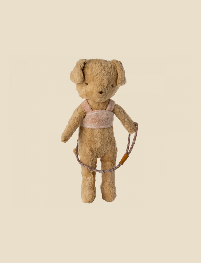 A well-worn vintage teddy bear stands upright, wearing a velvety harness that spells out "Maileg Puppy Supply, Harness" in cursive. The bear is fuzzy, with a light brown color and floppy ears. The background is light beige and empty.