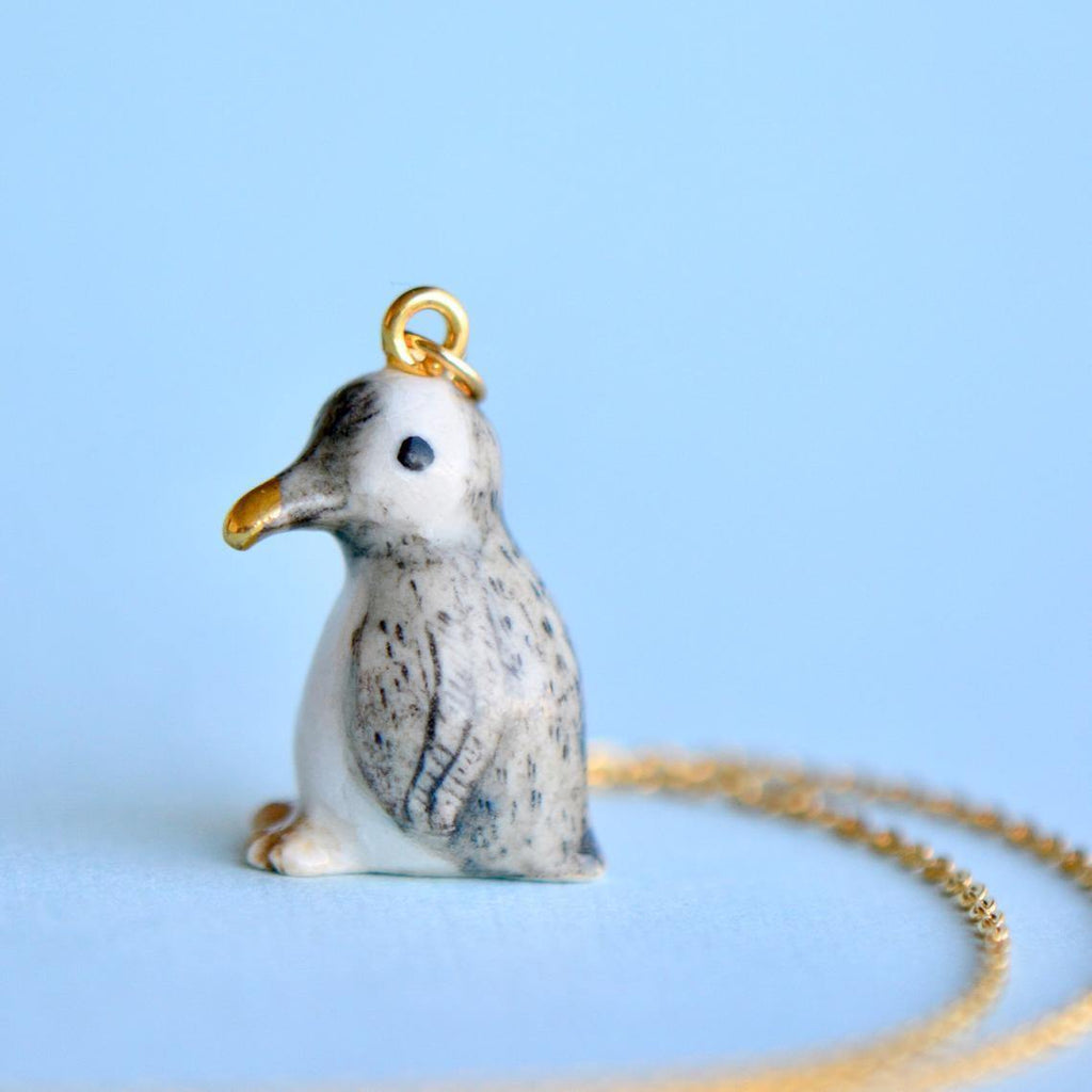 A close-up of the Tiny Penguin Necklace with a 24k gold plated ring on top, attached to a 24k gold plated chain necklace. The handmade jewelry piece features detailed grey and white coloring with a small black eye and a yellow beak. The background is a solid light blue.