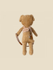 A plush teddy bear viewed from behind, featuring light brown fur and a cozy Maileg Puppy Supply, Harness with various miniature tools or accessories attached. The background is plain and light-colored, highlighting the teddy bear prominently.