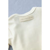 Close-up of the back of a cream-colored, organic cotton Baby Jumpsuit with a beige label sewn onto the neckline. The label reads "FOG LINEN WORK." The garment is displayed against a neutral background.