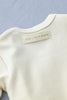 Close-up of the back of a cream-colored Baby Cap & Cardigan with a fabric tag that reads "FOG LINEN WORK." The tag is stitched into the neckline, and the garment appears to be made of soft, comfortable organic cotton. The background is a light gray surface.